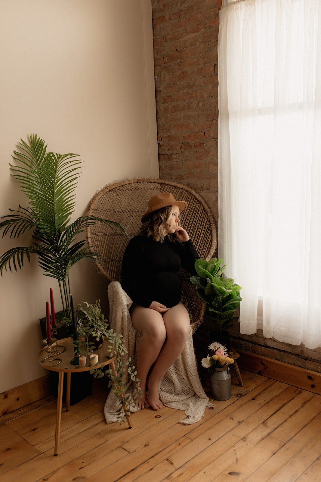 A mom to be sits in a large wicker chair surrounded by candles and plants looking out a large window