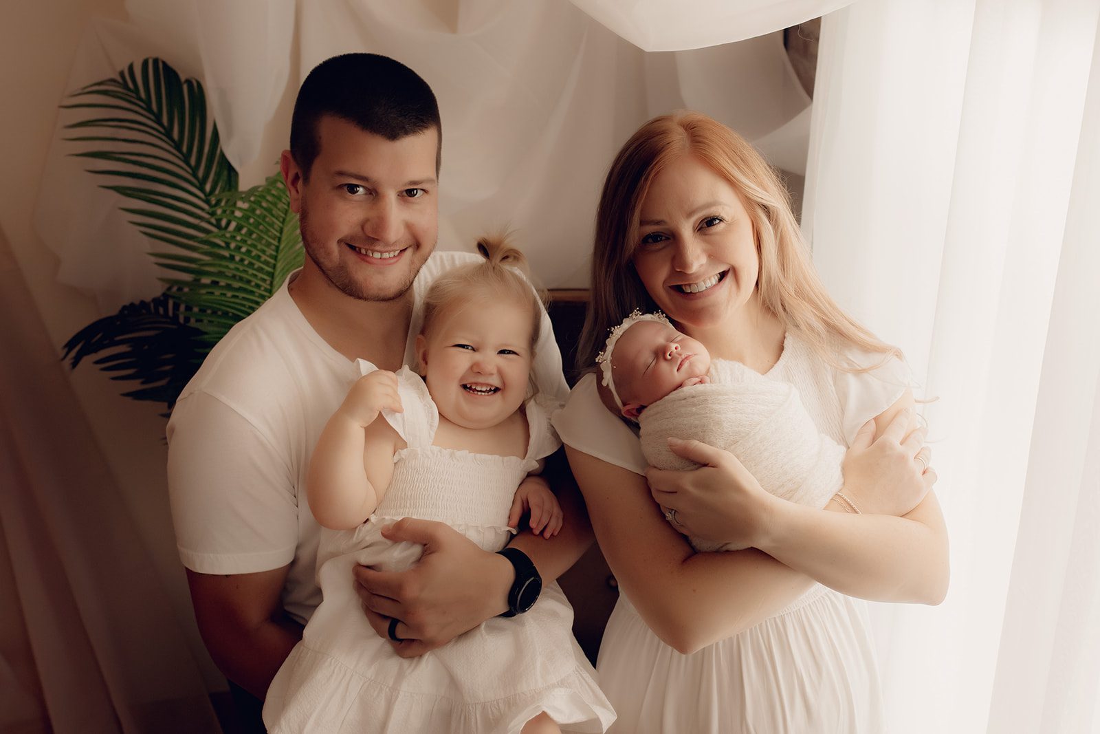 A mom in a white dress holds her sleeping newborn baby while dad holds their young toddler daughter in a white dress next to her after getting help from Beautiful birth choices