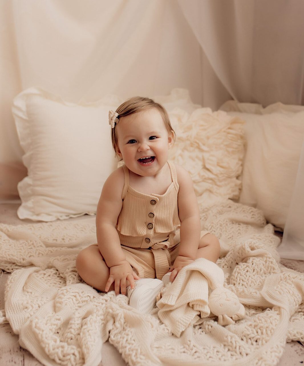 A toddler girl sits on a white bed laughing in a studio