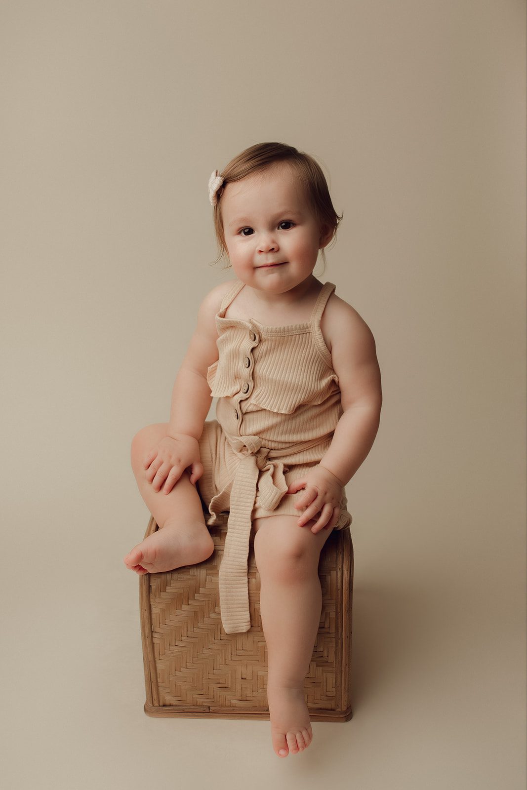 A toddler girl in a beige dress sits on a wicker box in a studio after visiting Healthy Baby Network