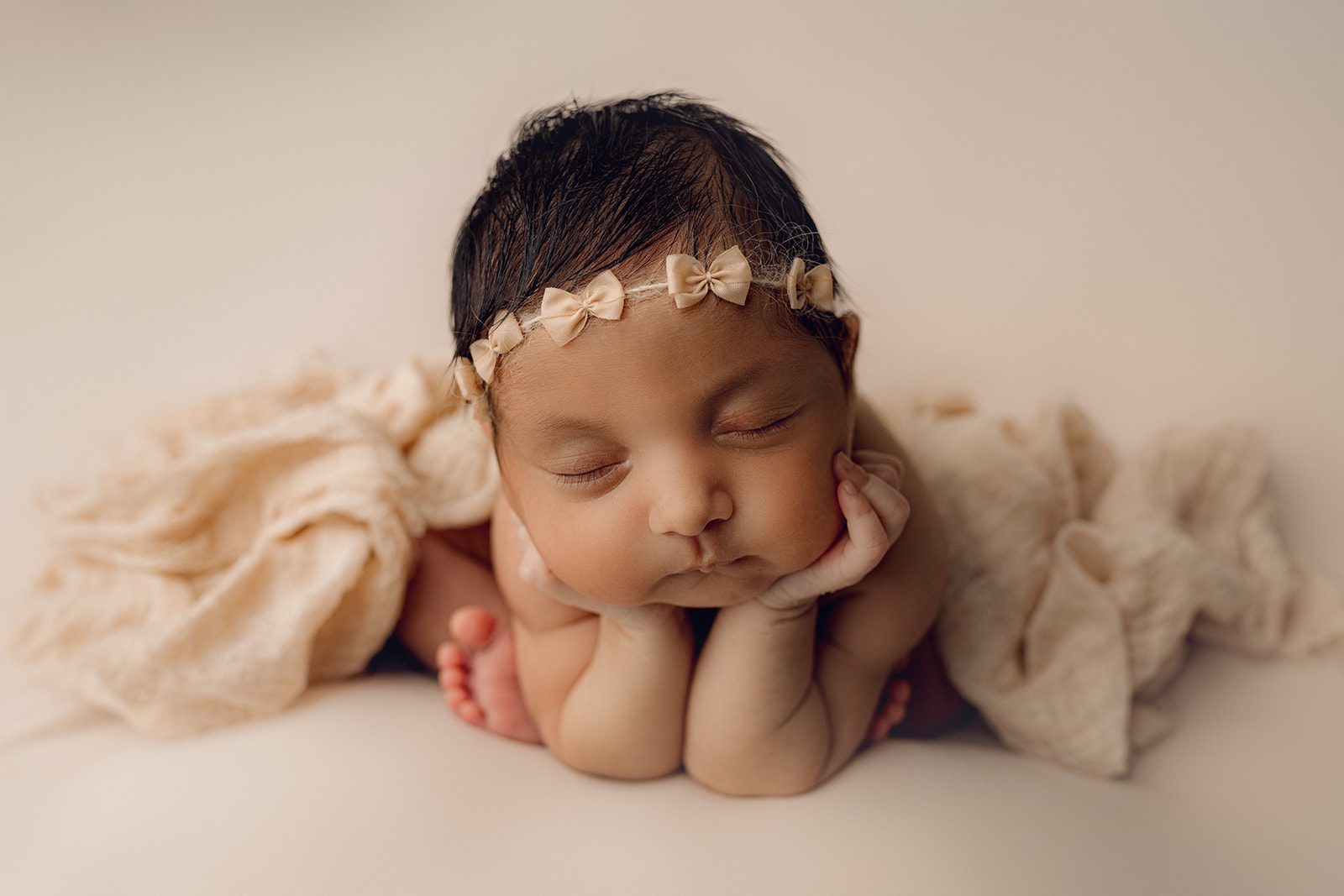 A newborn baby rests its head on its hands while wearing a beige bow headband