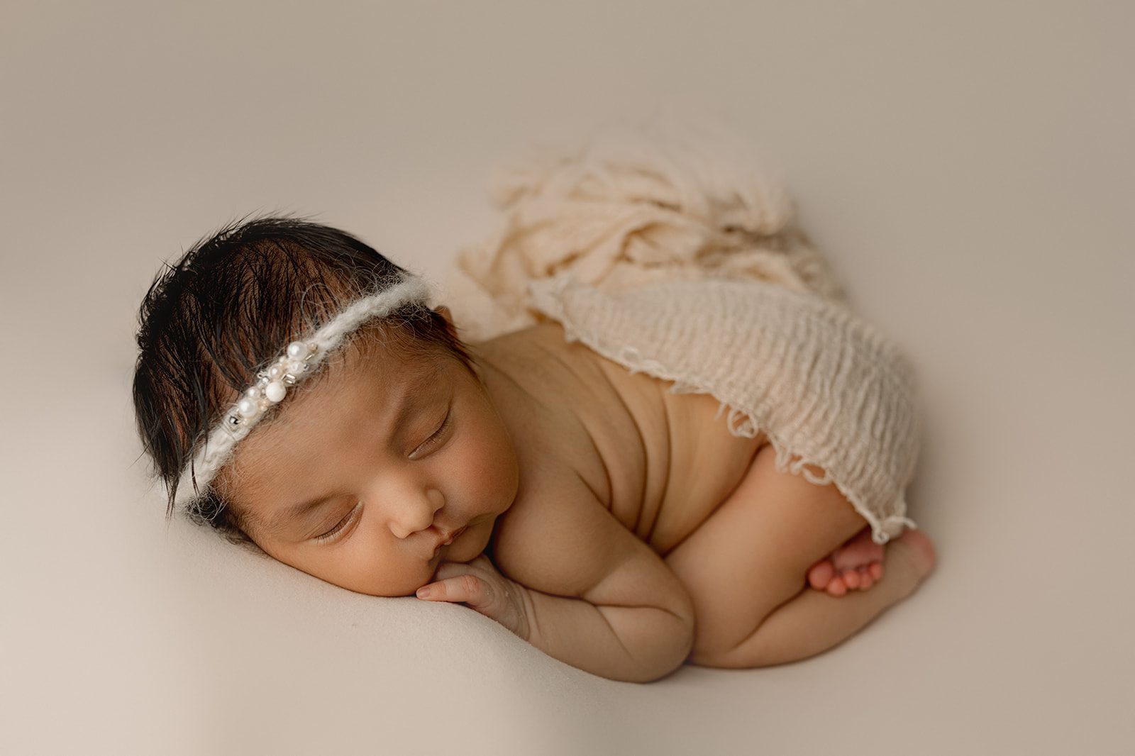 A newborn baby sleeps on its stomach under a beige blanket with a headband from Joyful Birth