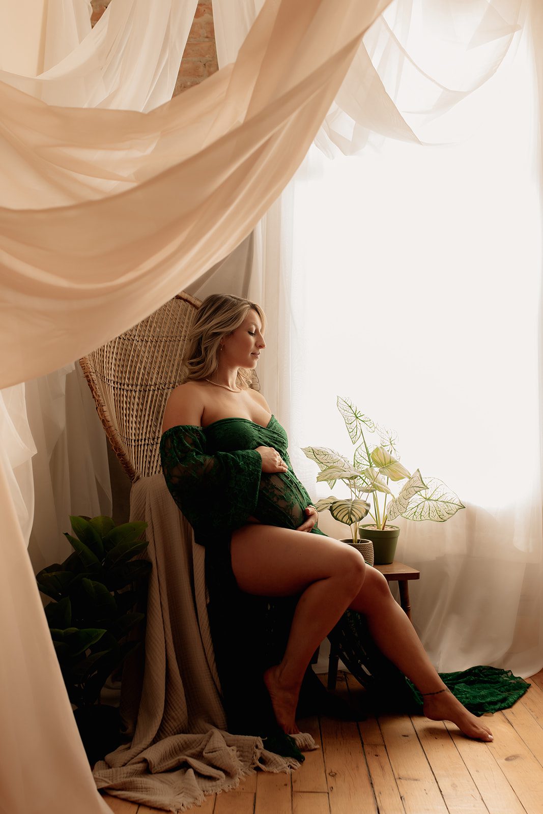 A mom to be leans back in a chair by a window while wearing a lace green maternity gown