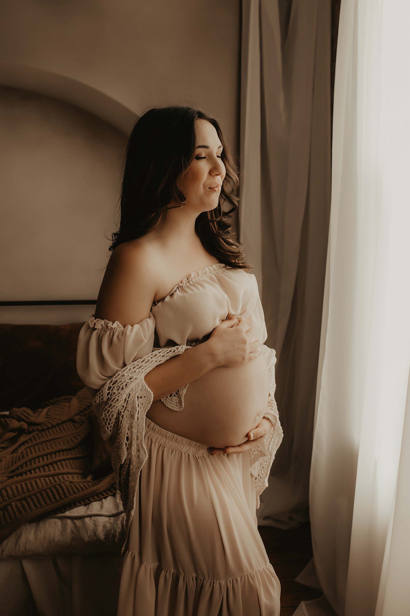 A mom to be smiles while standing in a window in a white skirt and crop top with hands on bump after visiting a birthing center in Rochester, NY