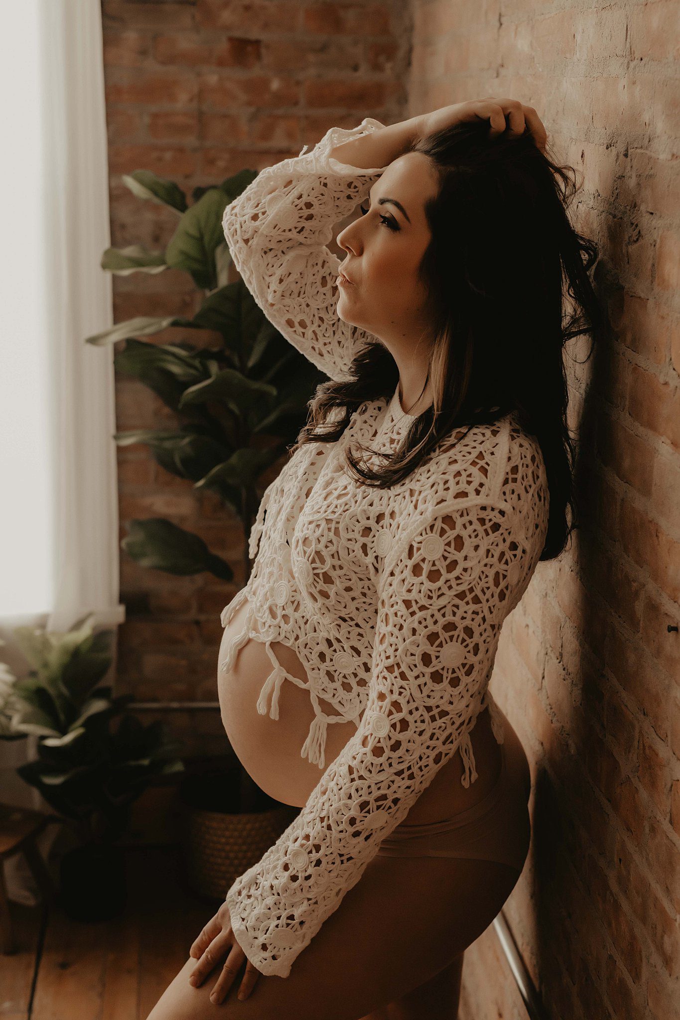 A mom to be leans against a brick wall in a lace top with a hand in her hair after visiting a birthing center in Rochester, NY