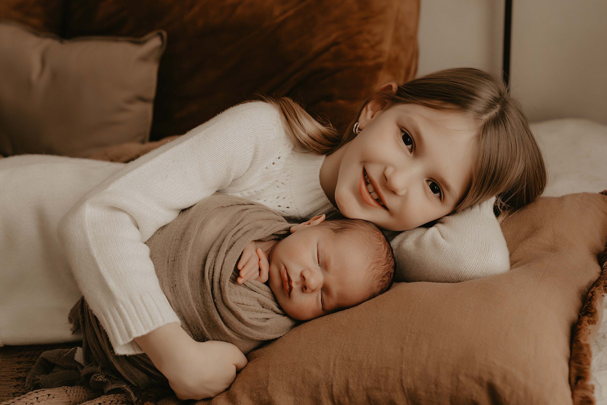 A young girl snuggles her sleeping newborn baby sibling on a bed in a white dress