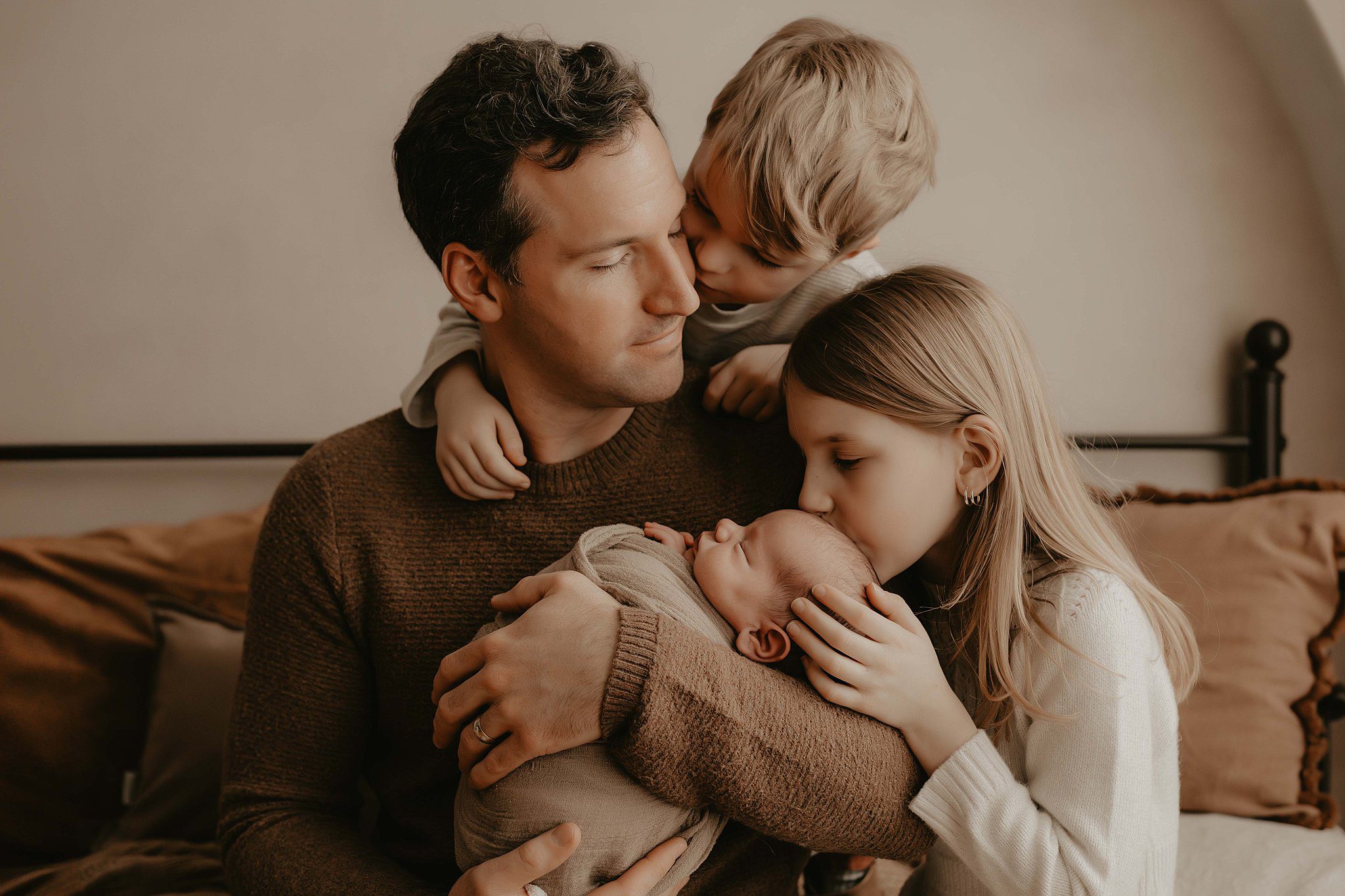 A dad holds his newborn as his young daughter and son kiss the baby and crawl on his shoulders on a bed after meeting a doula in Rochester, NY