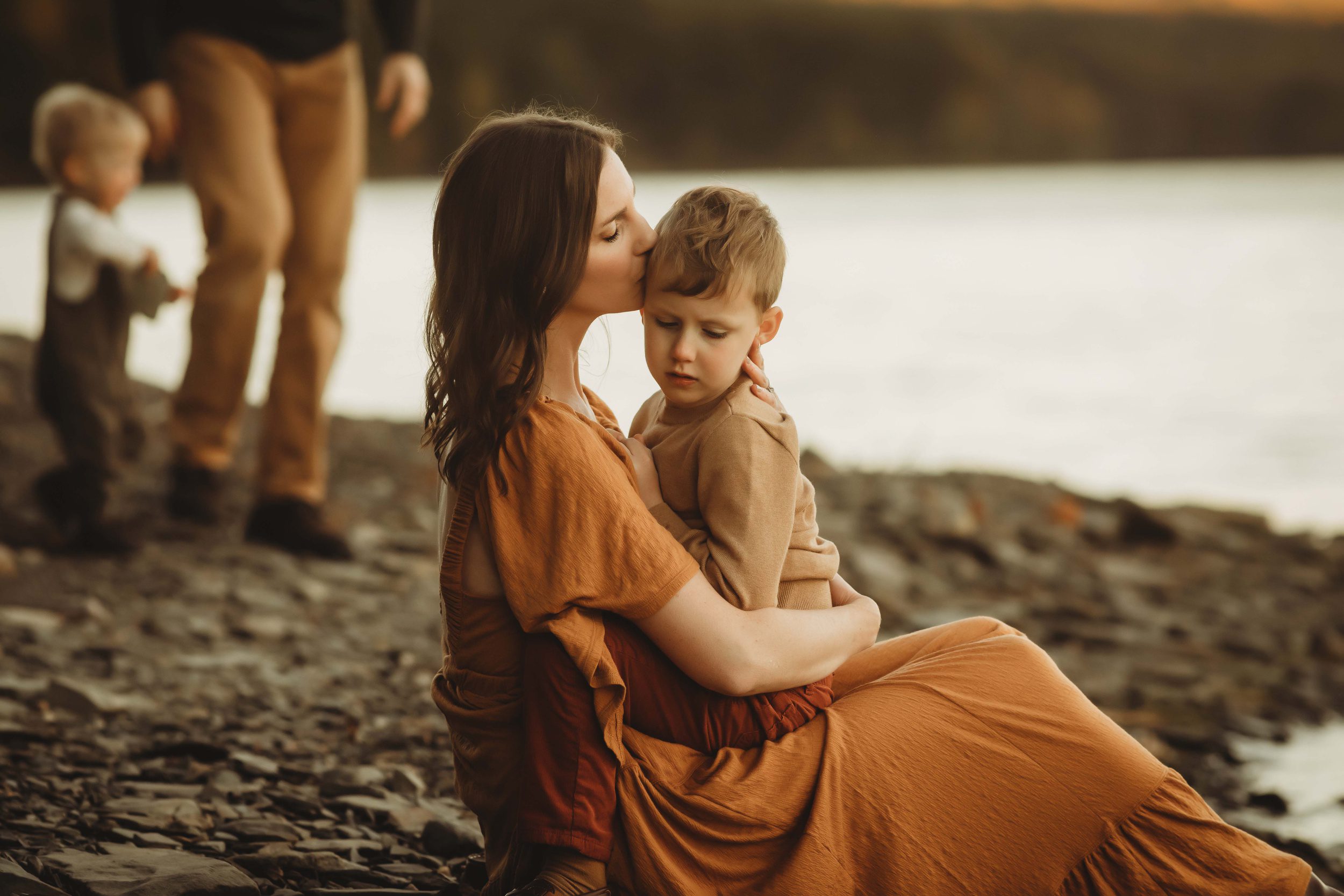 A mom kisses her young son in her lap while sitting on a beach at sunset in an orange dress after meeting nannies in rochester, ny