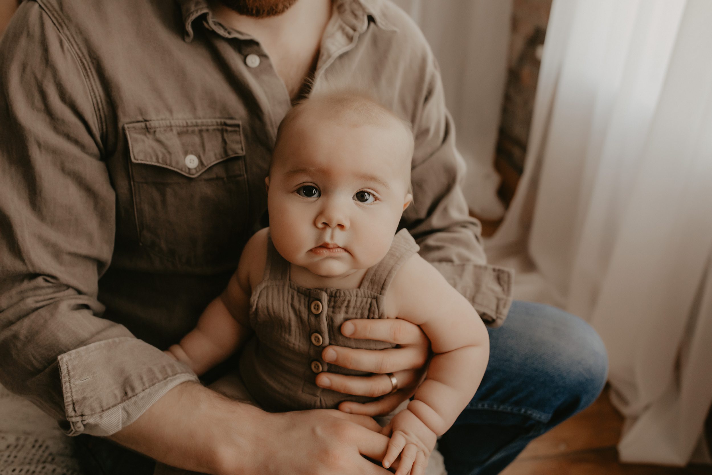 A baby sits in dad's lap on a bed under a window in brown overalls