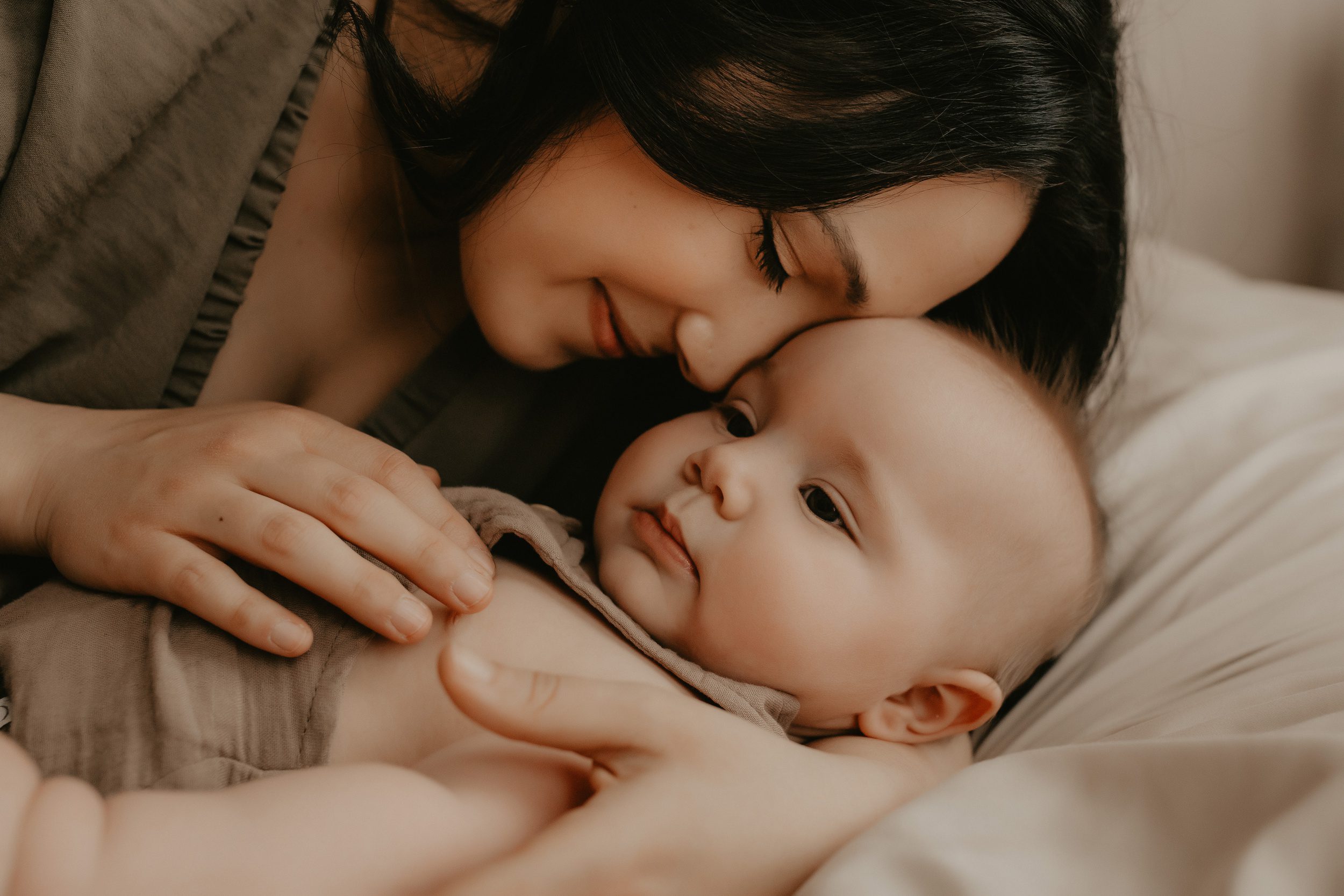 A happy mother on a bed snuggles with her newborn baby in brown overalls after meeting postpartum doulas in rochester, NY