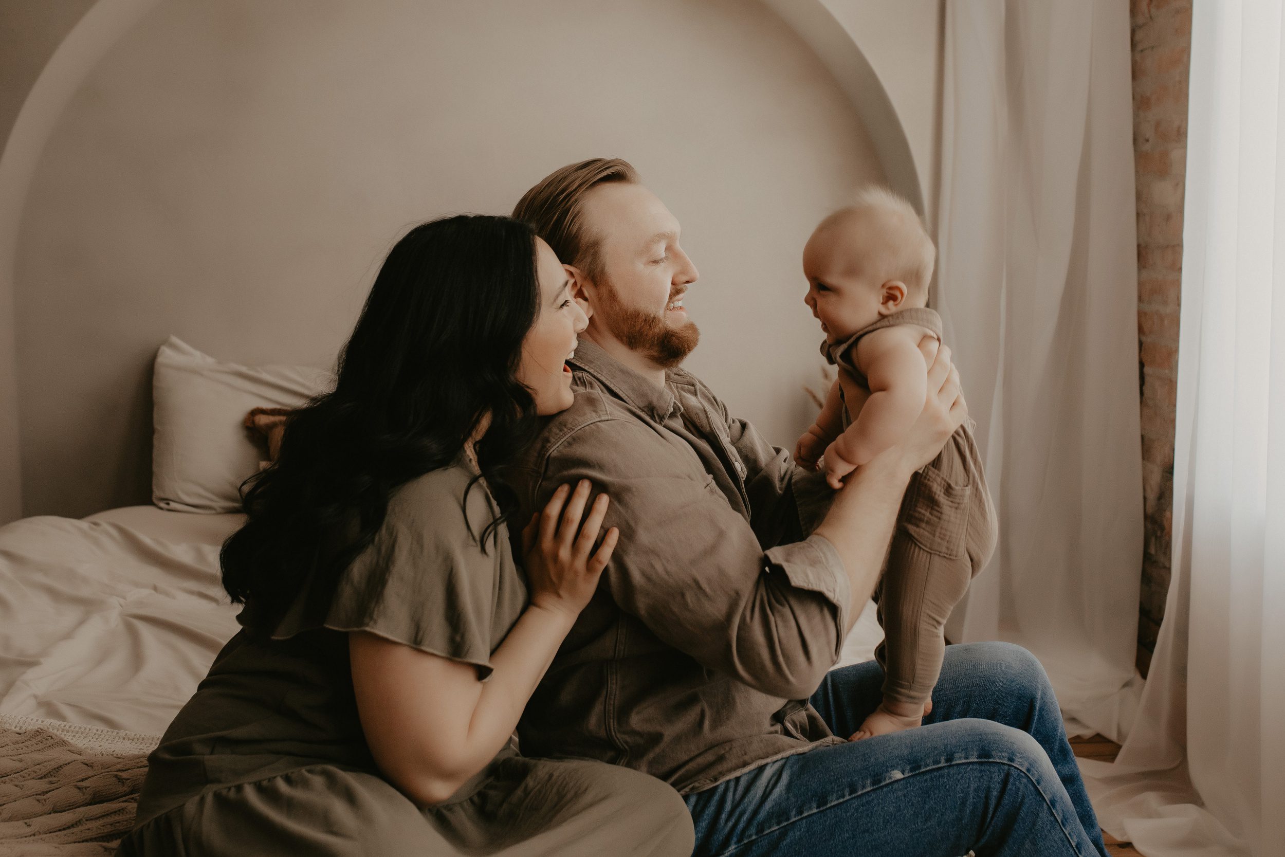 A mom and dad laugh with their baby on a bed in brown outfits after meeting postpartum doulas in rochester, NY